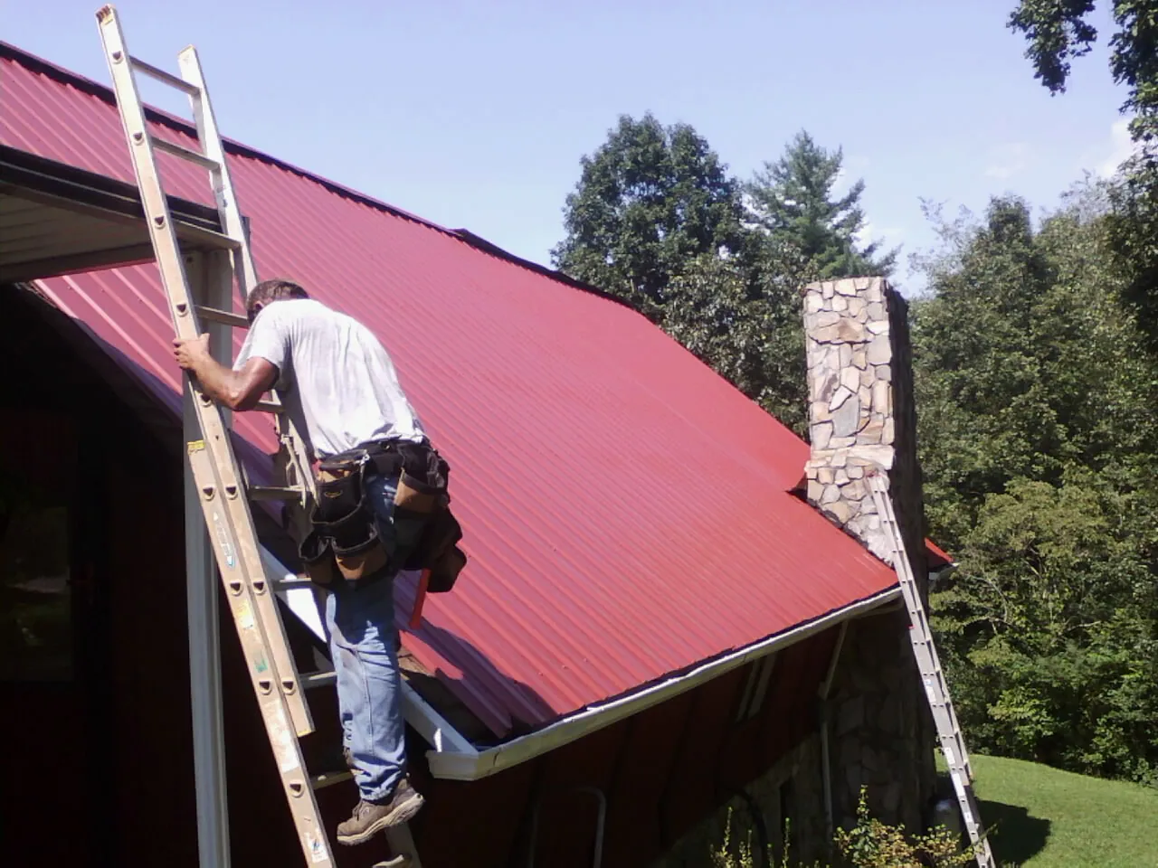 Roofer on a ladder beside a red metal roof and stone chimney — Soffit & Fascia Repair by Roofing Brothers in Tecumseh